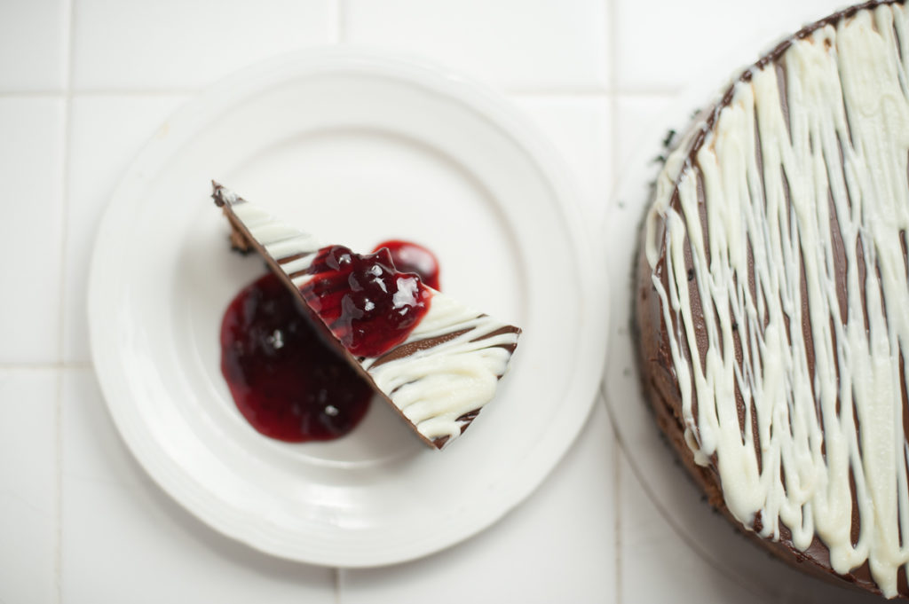 one slice of chocolate to the 4th power cheesecake on white plate garnished with Bonne Maman cherry preserves next to entire cheesecake