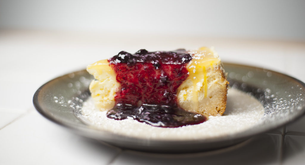 single slice of lemon lemon tart cheesecake garnished with blueberry preserves and confectioners sugar on dessert plate